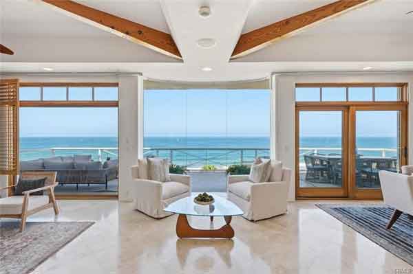 Bright beachfront living room with floor-to-ceiling ocean view, white sofas around a glass coffee table, and a glass balcony door.