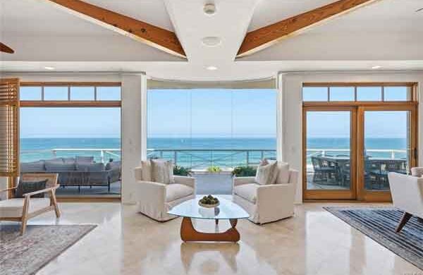 Bright coastal living room with white seating around a round glass coffee table, opening to a balcony with an ocean view.