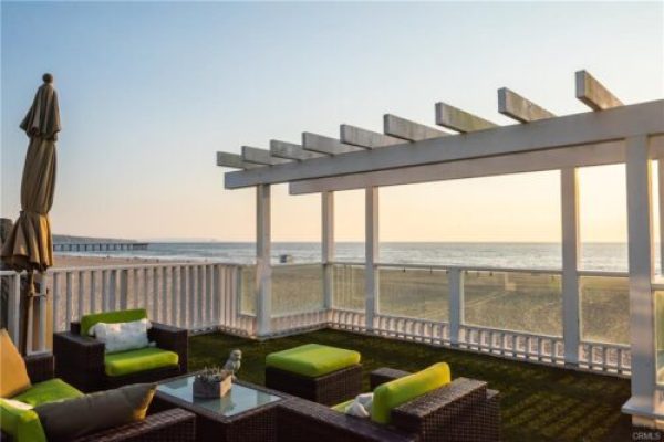 Outdoor deck seating with wicker chairs and lime-green cushions, overlooking the beach at sunset through a glass railing and pergola beams.